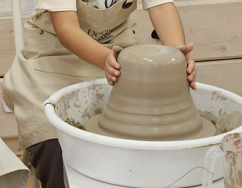 앞치마를 두르고 돌아가는 물레 위 흙에 두 손을 얹은 모습